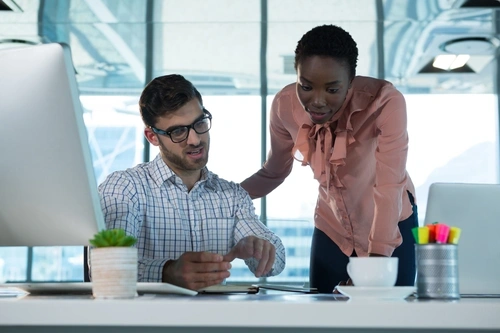 Professional female leaning over a male coworker's desk to review their work.